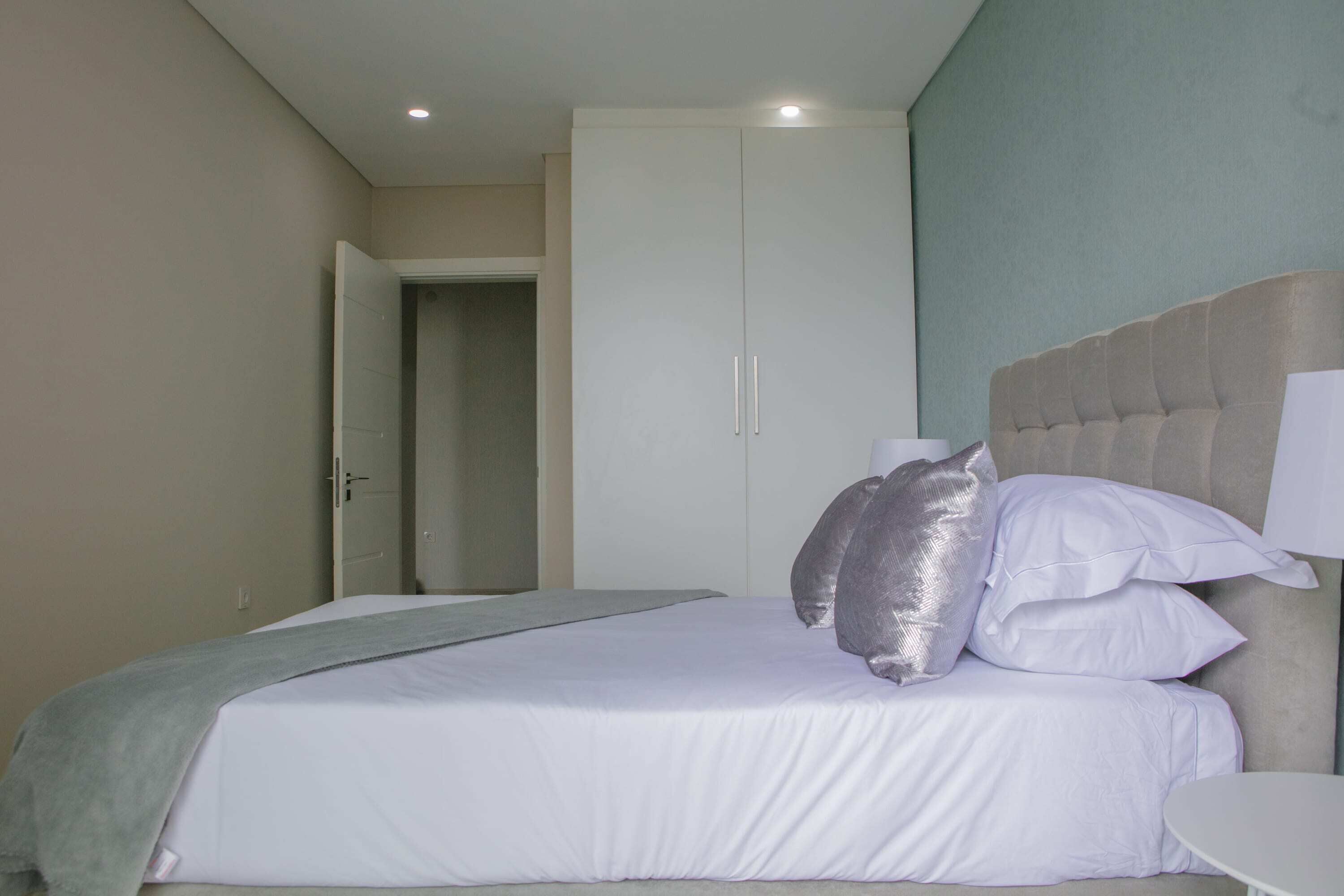 A bedroom with a bed, a bedside table, a wardrobe, and a door leading to another room.