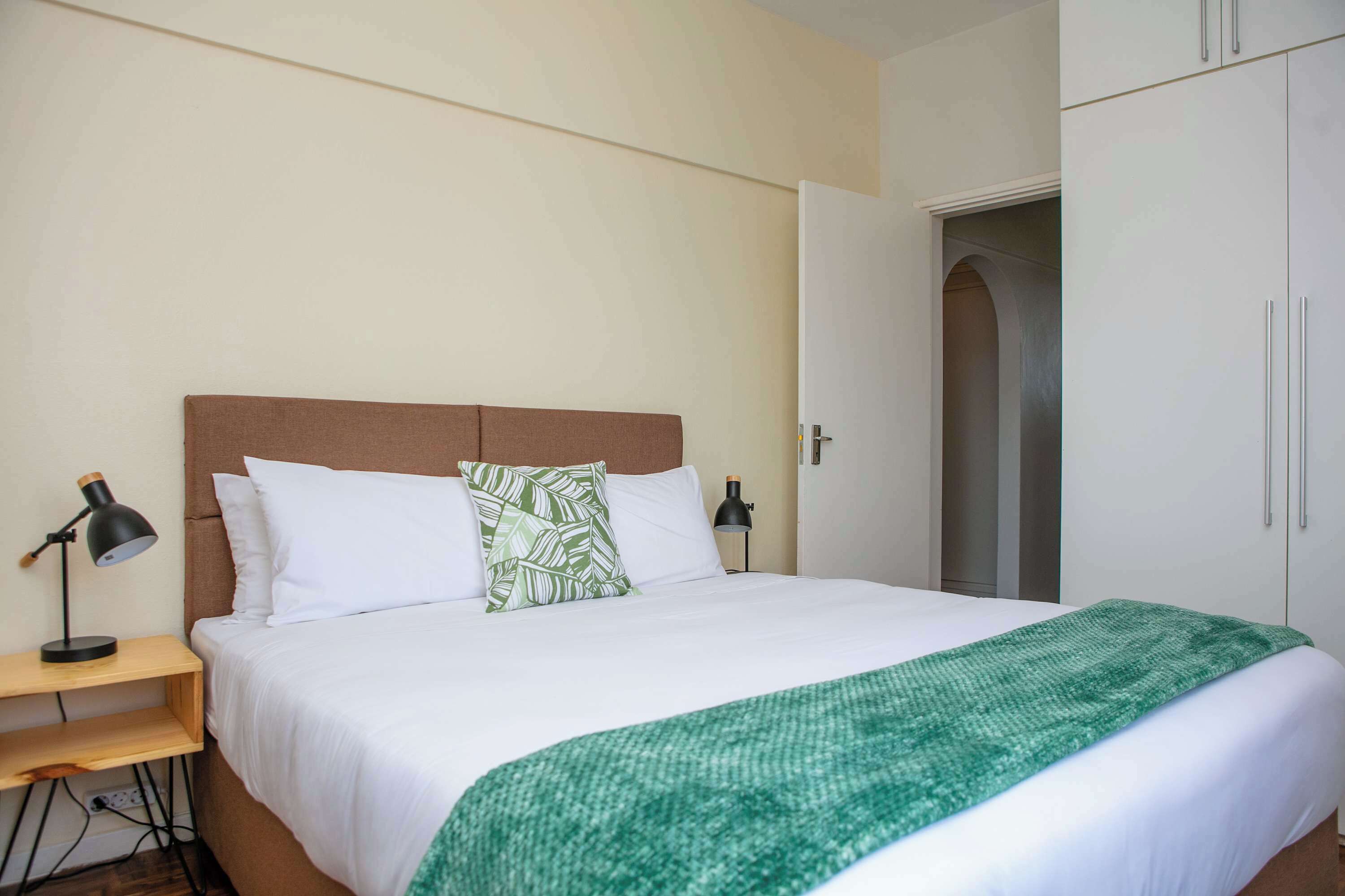 A neatly made bed with white bedding and a green throw blanket, a bedside table with a lamp, and a wardrobe with a door ajar.