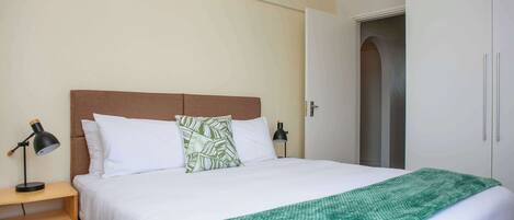 A neatly made bed with white bedding and a green throw blanket, a bedside table with a lamp, and a wardrobe with a door ajar.