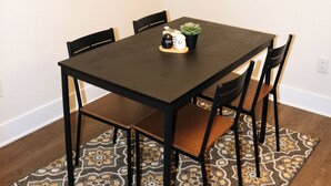 Un coin repas avec une table en bois foncé, quatre chaises, un tapis à motifs, une fenêtre avec des stores et une estampe botanique encadrée au mur.