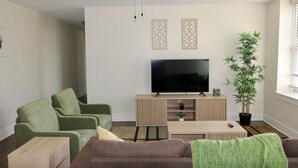 Un salon équipé d’un téléviseur à écran plat, de deux fauteuils verts, d’un canapé marron, d’une table basse en bois, d’un ventilateur de plafond et d’une plante en pot.