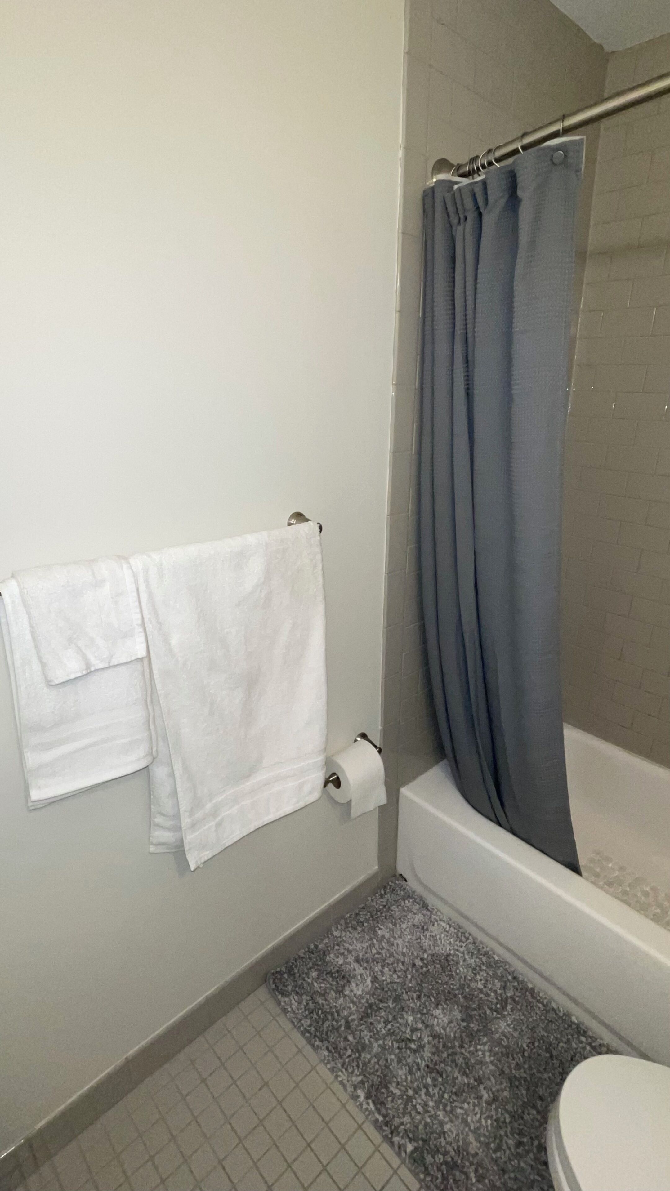 Et badeværelse med et hvidt badekar, en grå bruseforhæng, et hvidt håndklæde på en holder og en toiletpapirholder.
