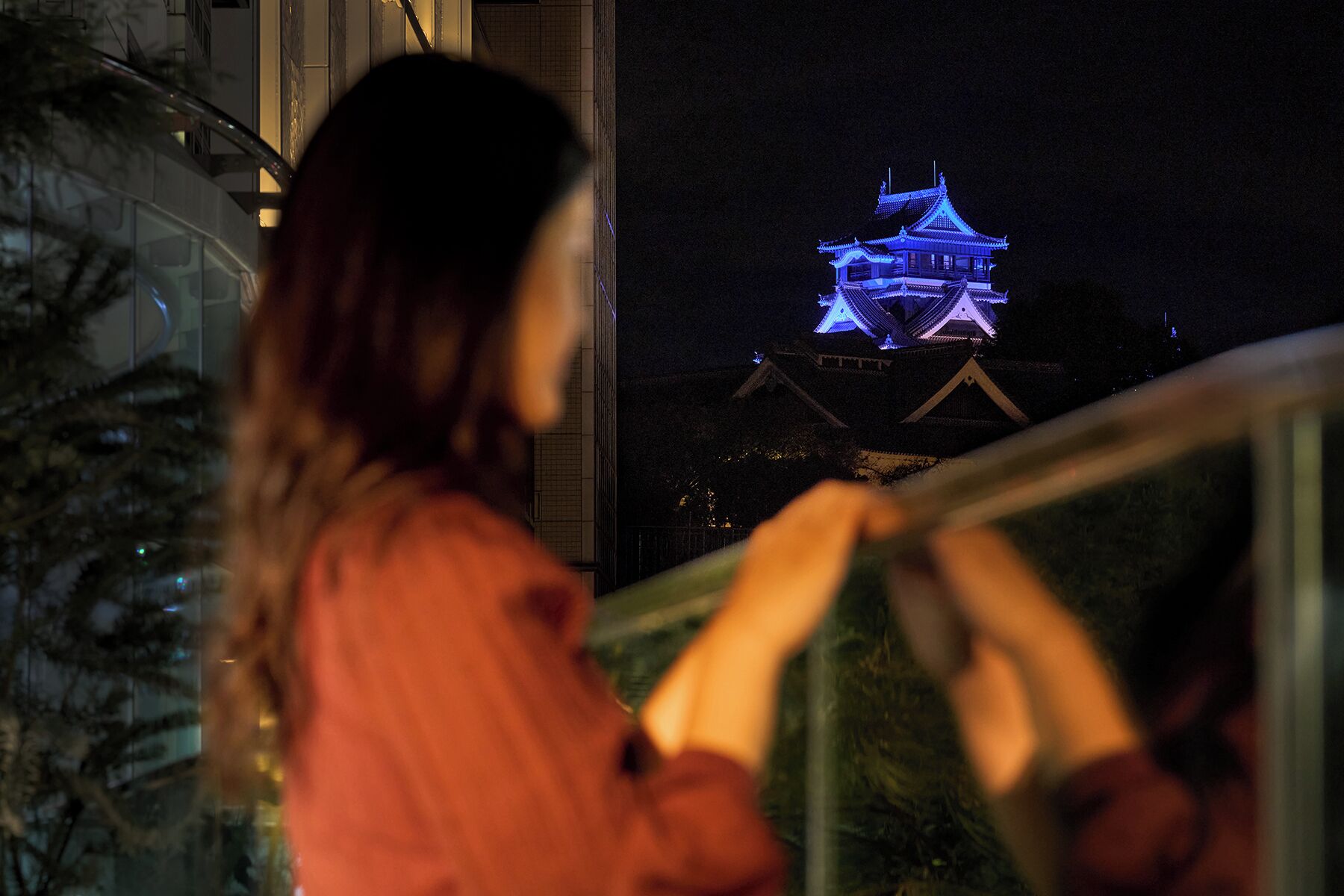 En person står ved et gelænder og ser ud over en traditionel bygning oplyst i blåt om natten.