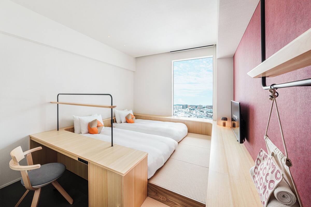 Habitación de hotel moderna con un escritorio de madera, dos camas, una silla, un televisor y vistas a la ciudad.
