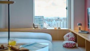 Habitación de hotel con cama, escritorio con lámpara, ventana con vista a la ciudad y un pequeño rincón de descanso con cojines.