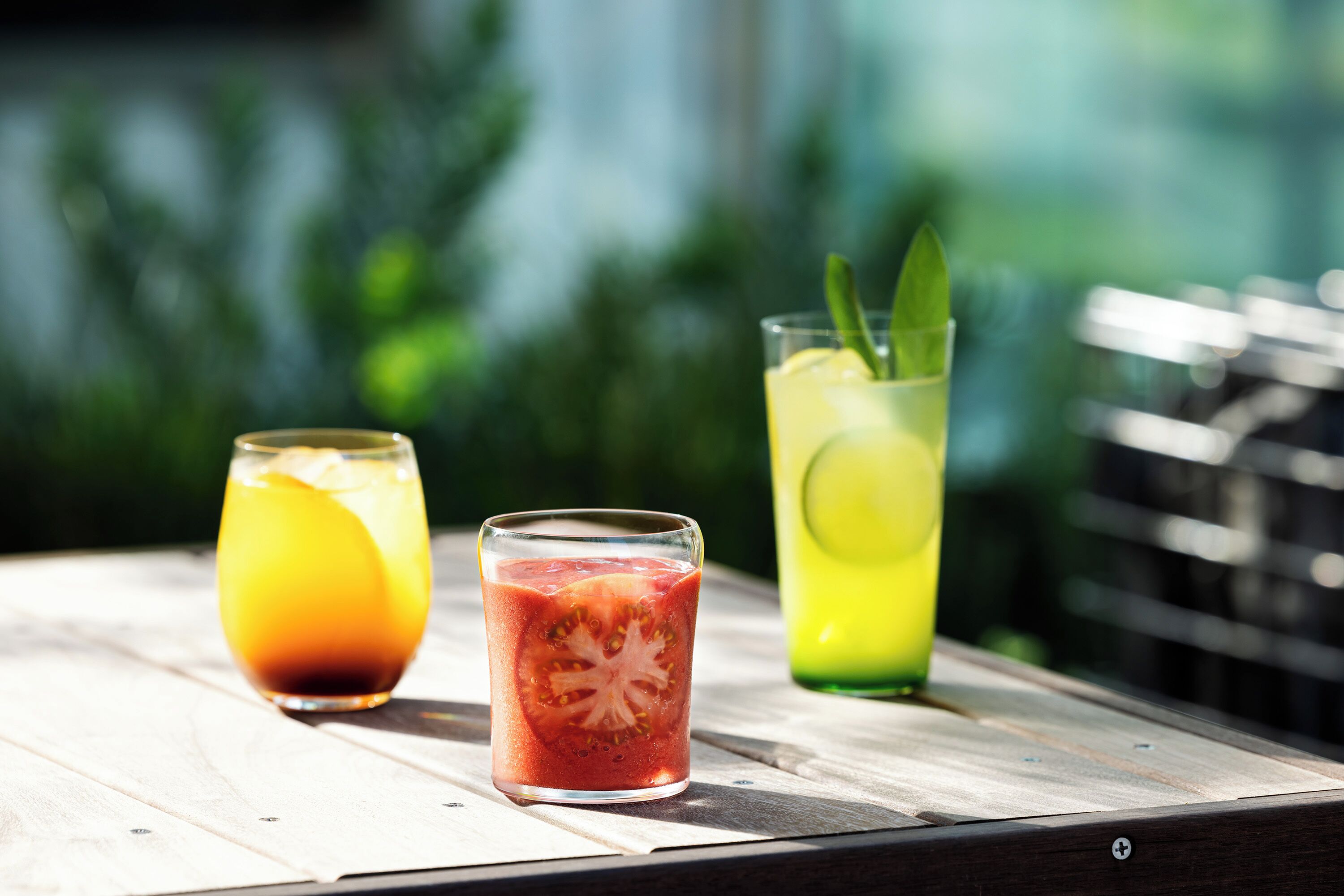 Three glasses of beverages on a wooden table with a blurred green background.