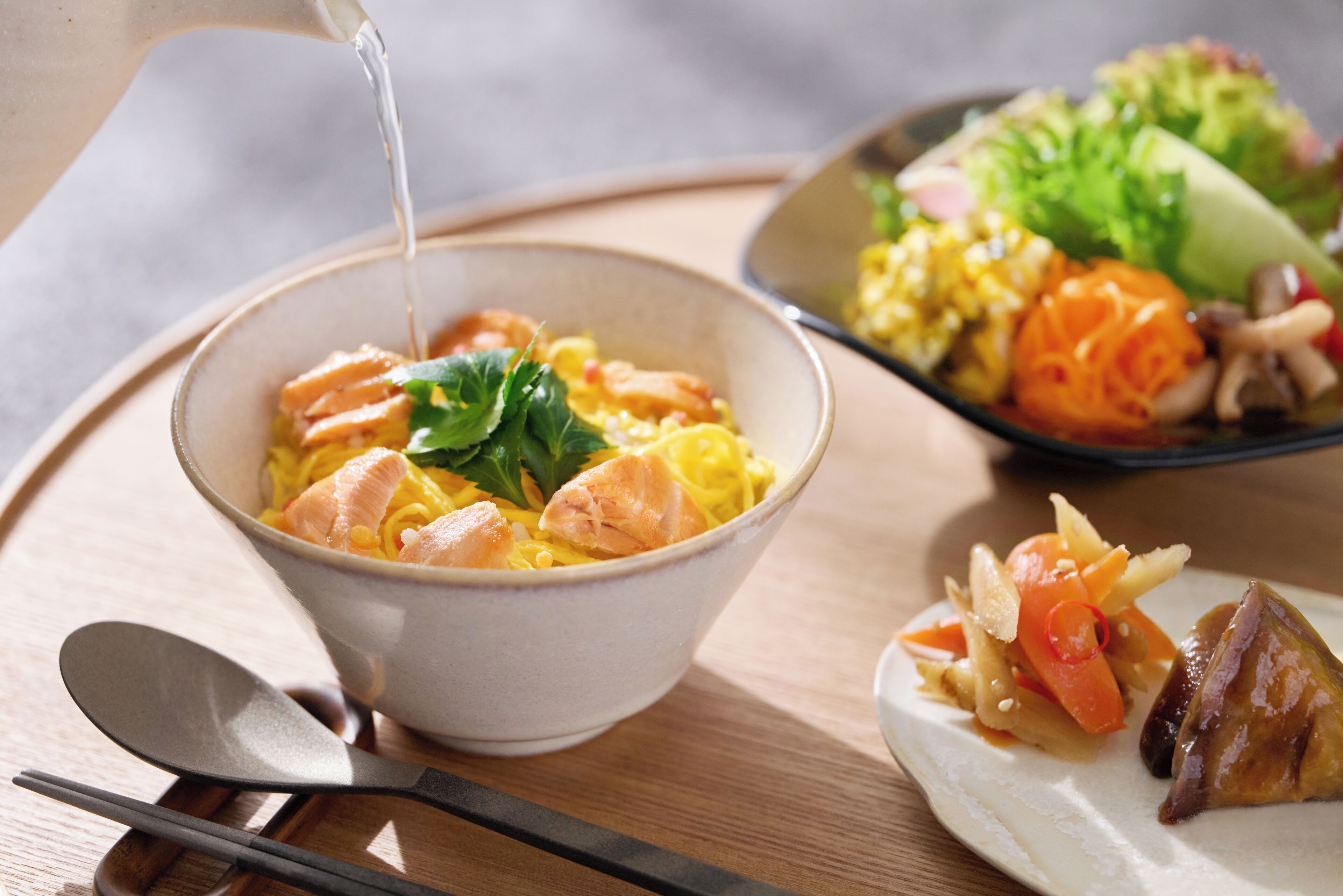 Eine Schüssel Ramen mit Lachs und Gemüse, ein Salat und ein Fleischstück auf einer Holztischplatte.