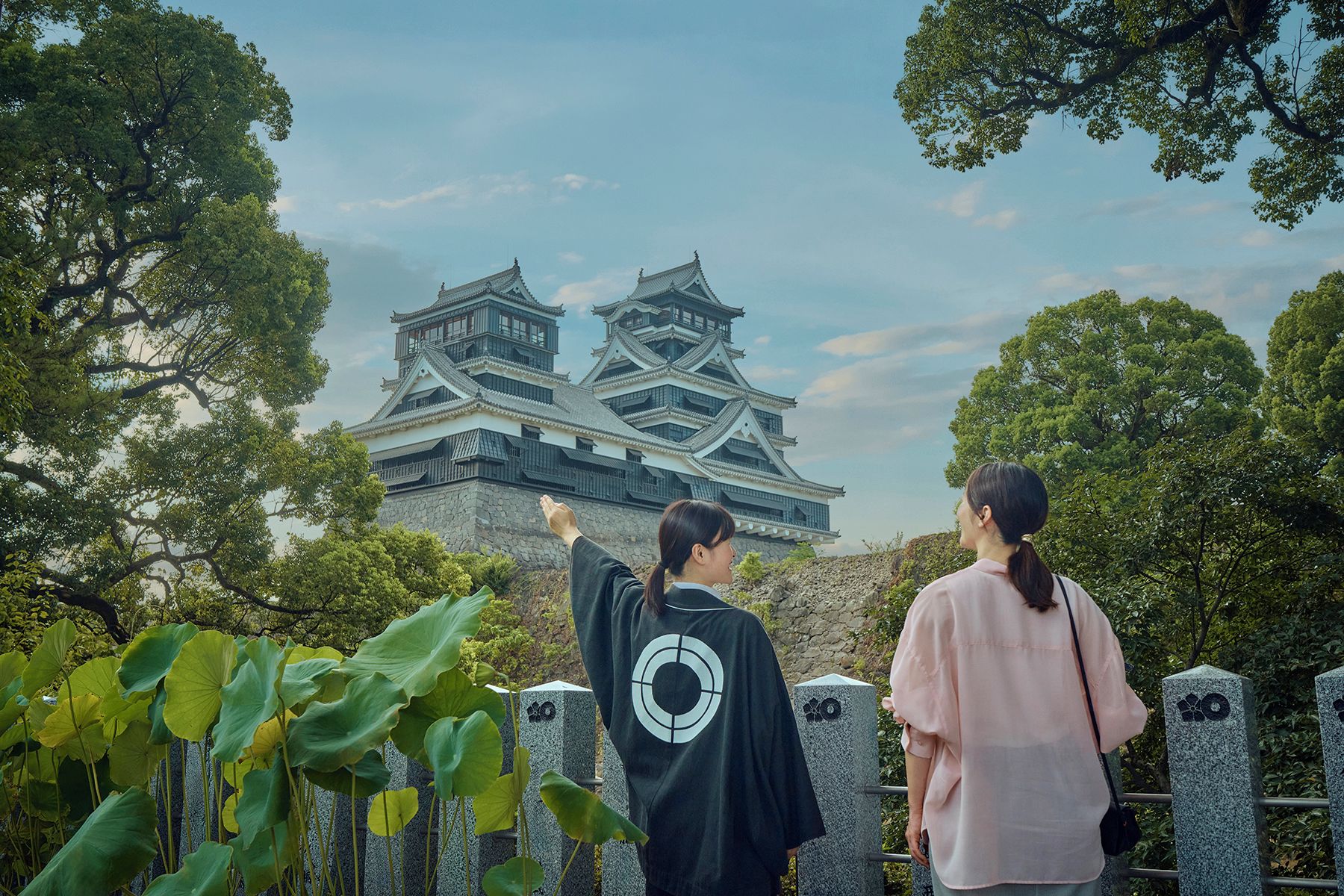 Two individuals observing a traditional Japanese castle with multiple tiers and a stone wall in the foreground.