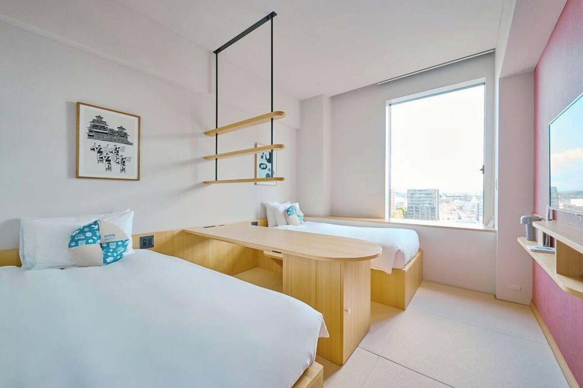 Habitación de hotel con dos camas, un escritorio de madera, un ventanal con vistas a la ciudad y una obra de arte enmarcada en la pared.