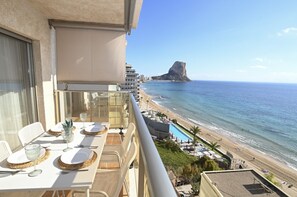 Ein Balkon mit Esstisch, Blick auf einen Strand mit einer markanten Felsformation, einen Pool und einen klaren Himmel.