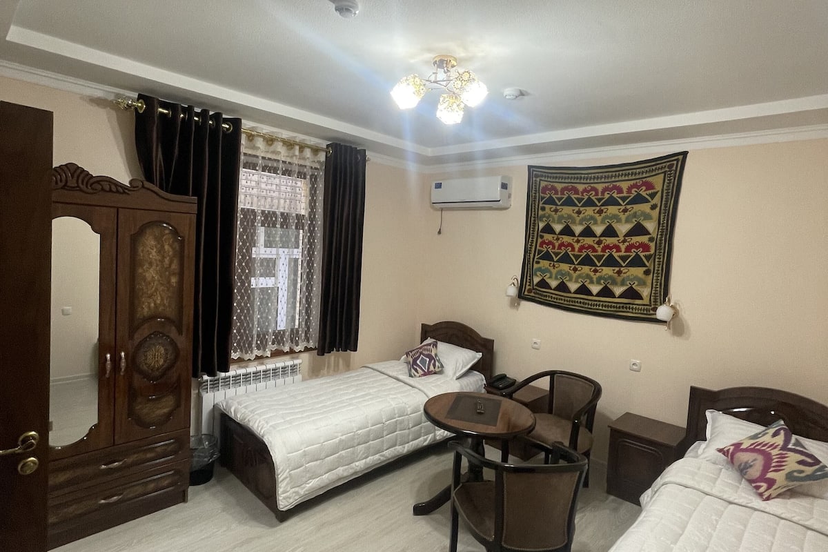 A hotel room with two single beds, a wooden wardrobe, a desk with a chair, a small table, and a decorative wall hanging.