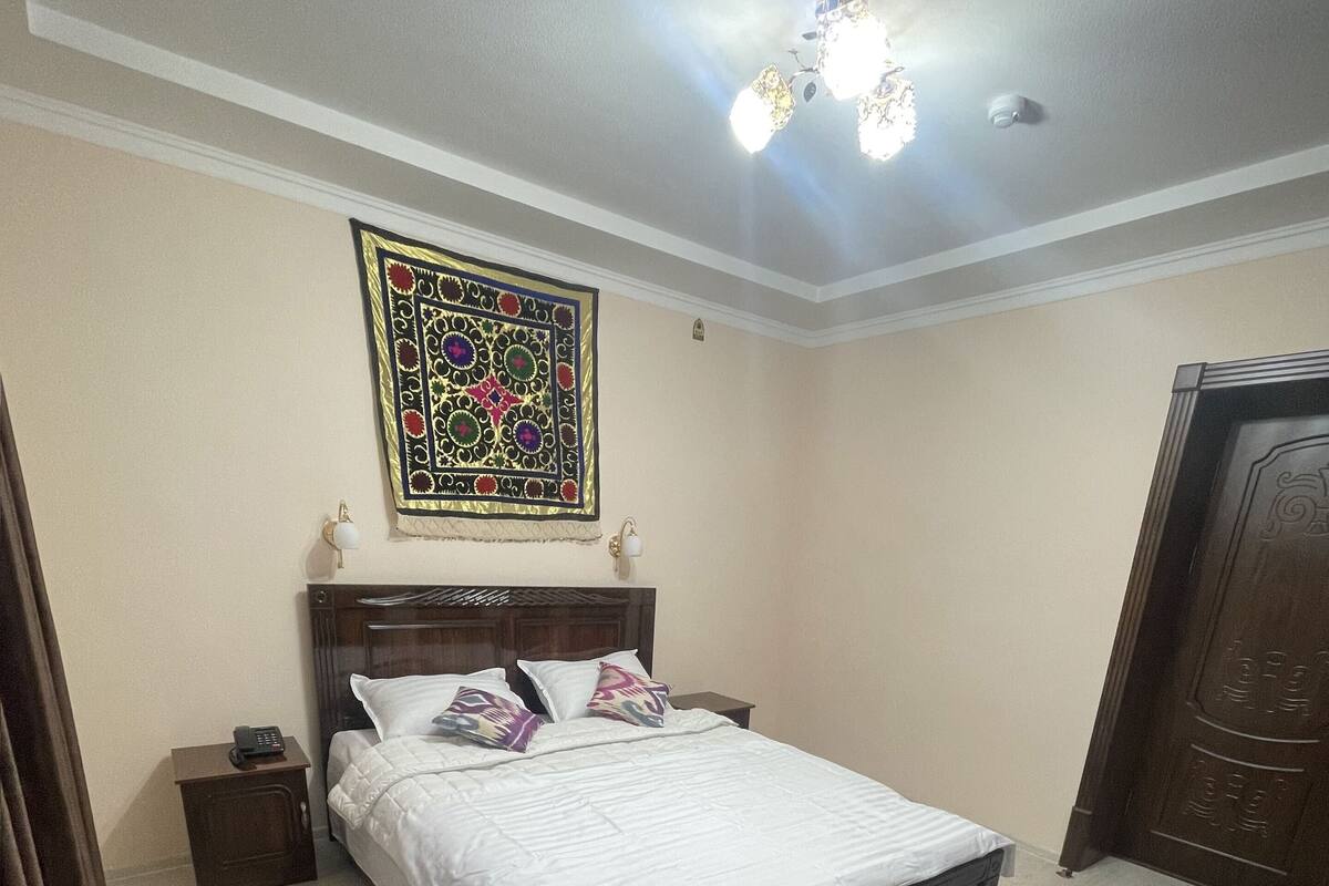 A hotel room with a bed, bedside table, lamp, and a decorative wall hanging.