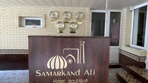 Una mesa de recepción con un cartel que dice "Samarkand Ali Hotel boutique" y una pared con relojes que marcan diferentes ciudades.