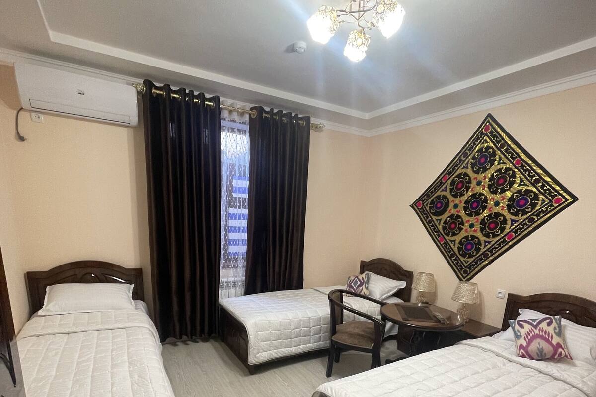 A hotel room with three single beds, a small table, a chair, a decorative wall hanging, and an air conditioning unit.