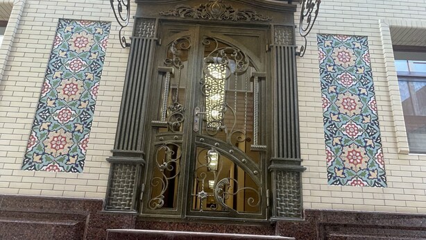 Una entrada de edificio con puertas de metal ornamentadas, un muro de azulejos con diseño y suelo de baldosas.