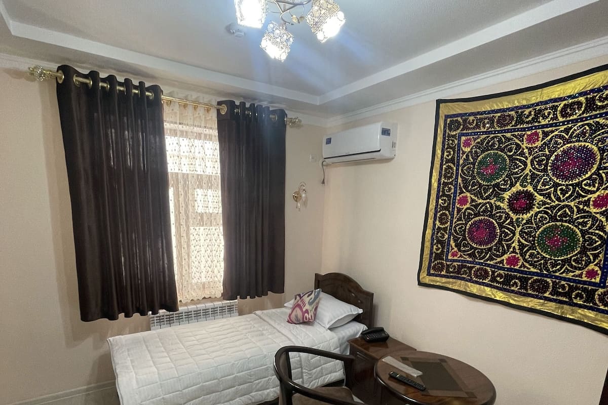 A hotel room with a bed, a desk, a chair, an air conditioner, and a decorative wall hanging.