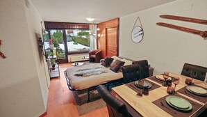 A compact room with a bed, a dining table set for four, and a view of the outdoors through a sliding door.