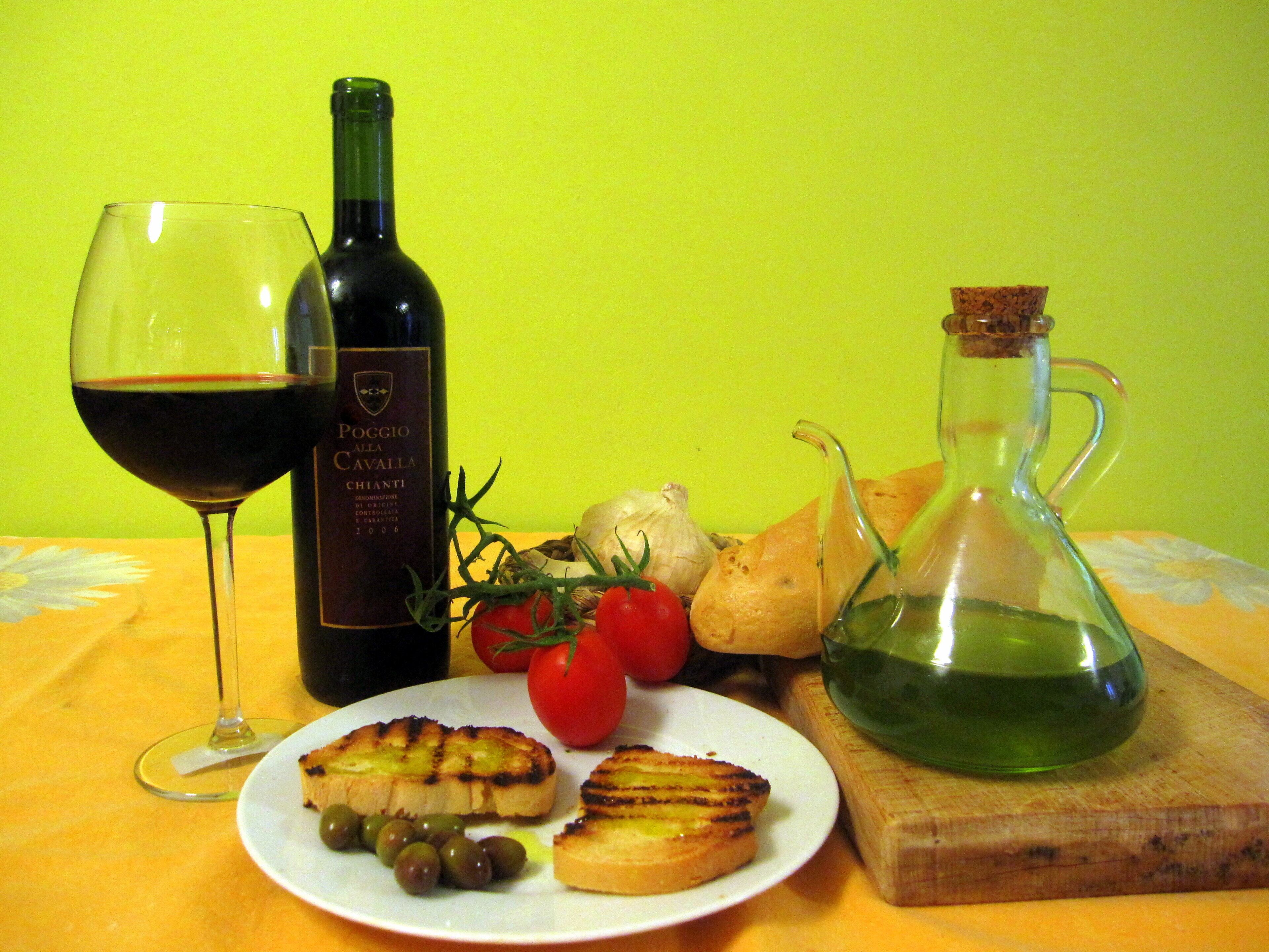 A plate with grilled cheese, tomatoes, and olives, a bottle of wine, and a jug of olive oil on a table.