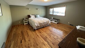 Un dormitorio con piso de madera, una cama con ropa blanca, una mesita de noche, una pequeña mesa y un ventanal con vistas.