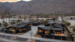 Een modern hotel met meerdere gebouwen met zwarte daken, gelegen in een binnentuin met palmbomen en een berg op de achtergrond.
