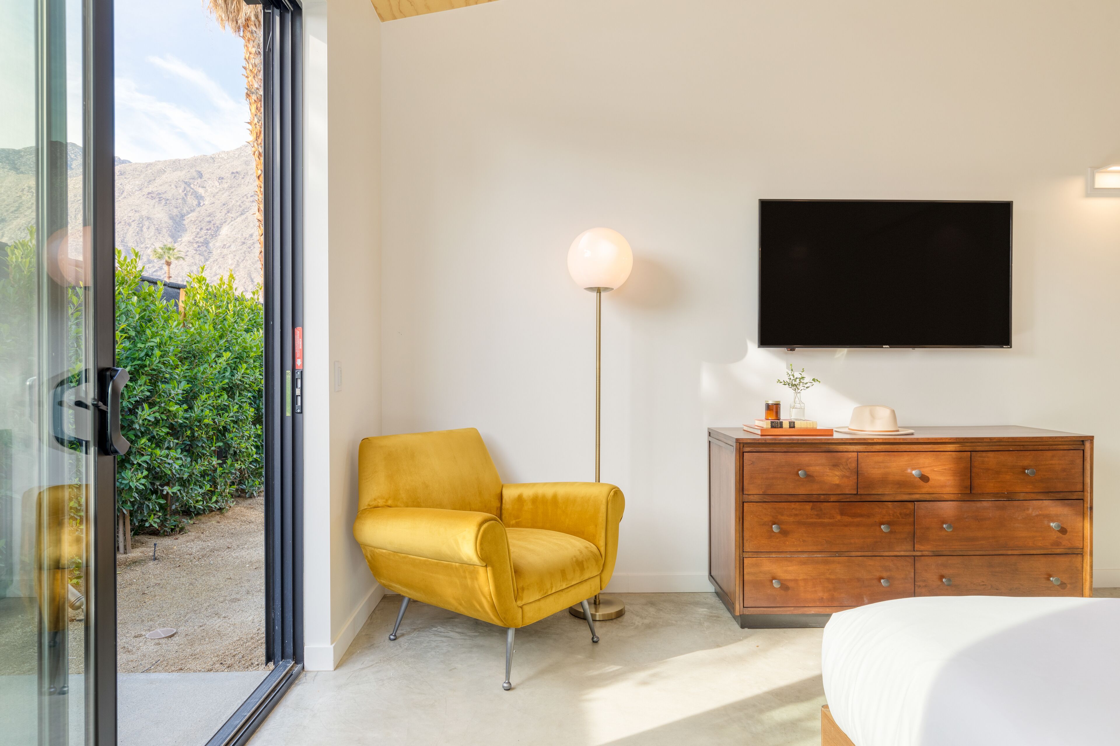 Ein Zimmer mit gelbem Sessel, einem Holzkasten, einem Flachbildfernseher und einem großen Fenster mit Blick auf Grünflächen und Berge.