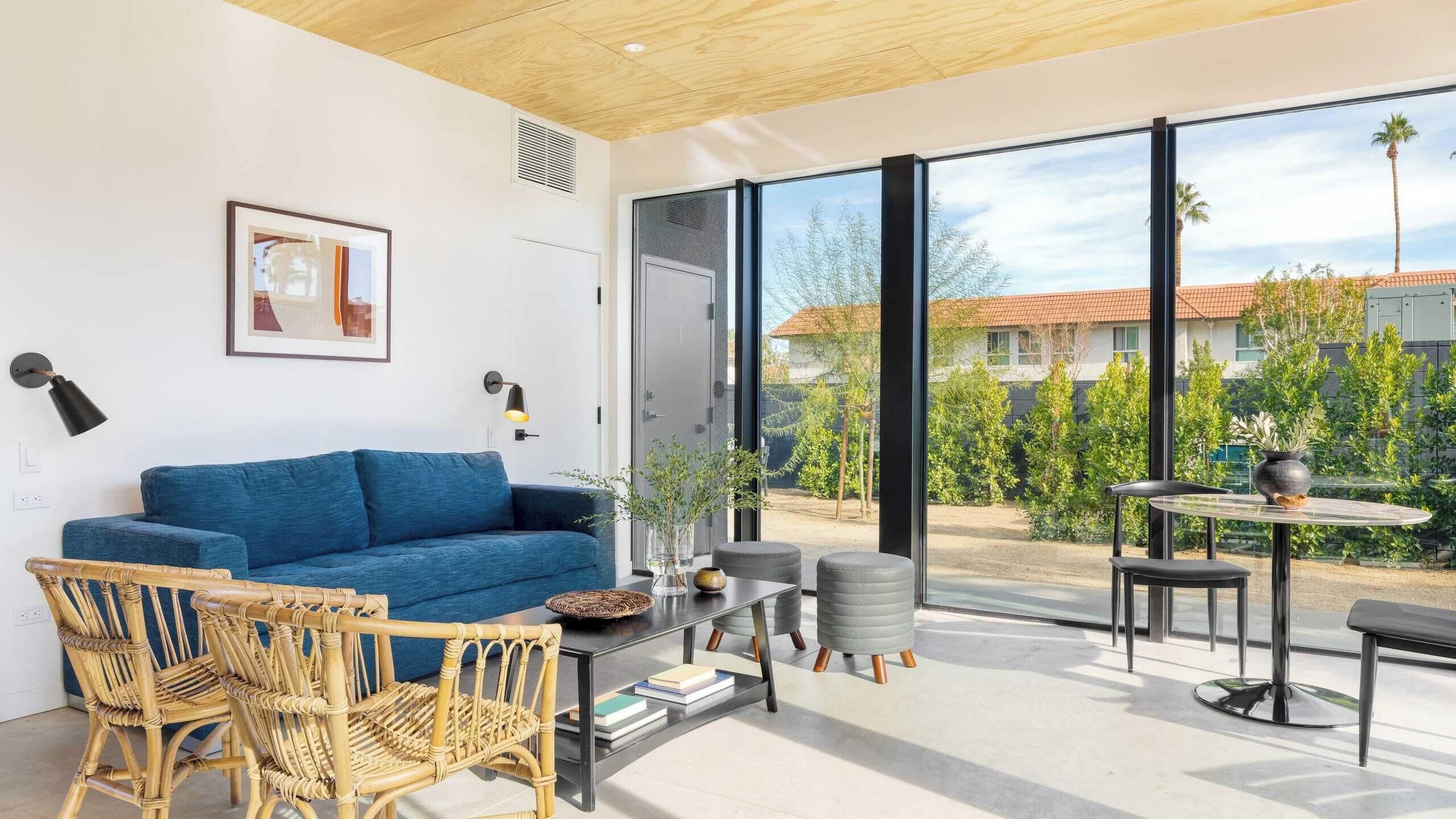 A modern living room with a blue sofa, wicker chairs, a coffee table, and a view of a garden and buildings through large windows.