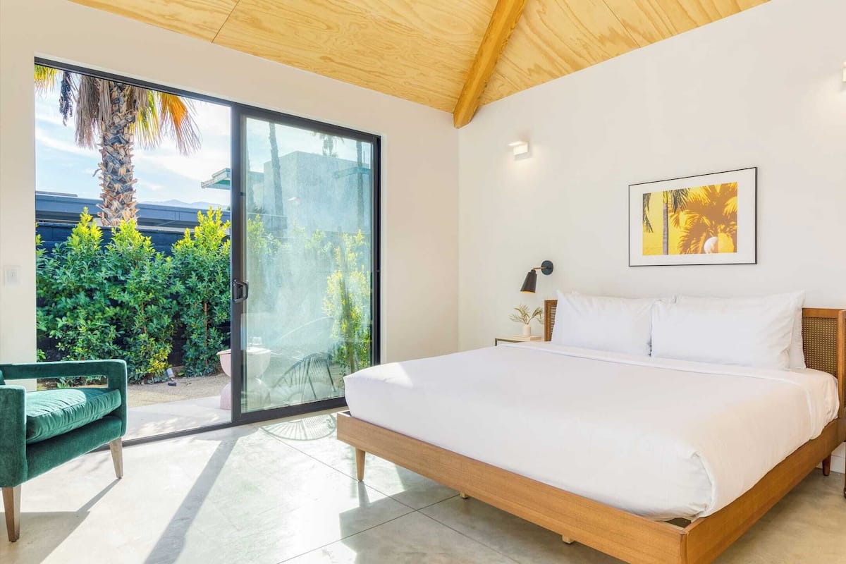 A modern bedroom with a large bed, a green chair, and a view of greenery through a sliding door.