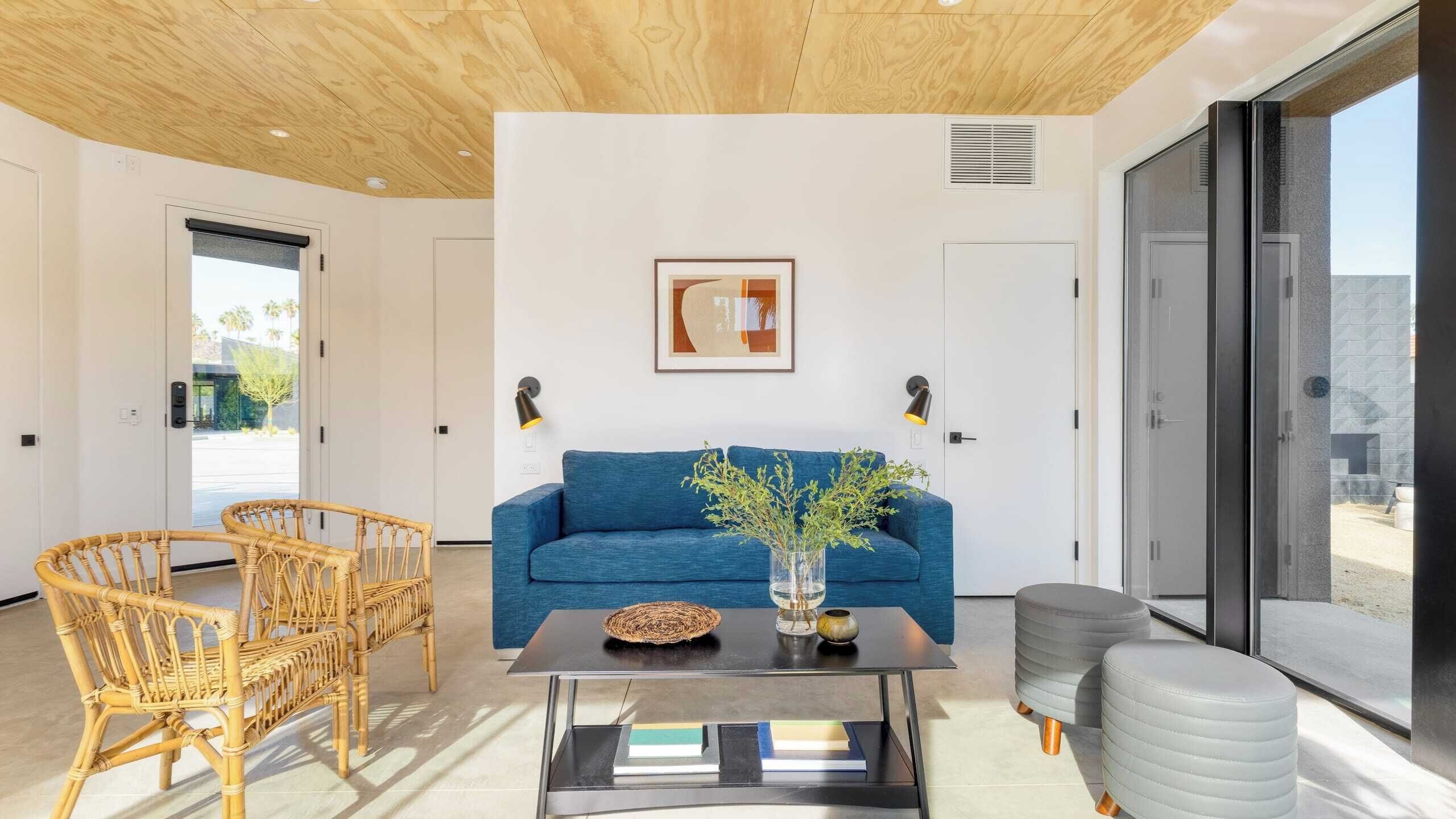 A modern living room with a blue sofa, wicker chairs, a coffee table, and a large window offering a view.