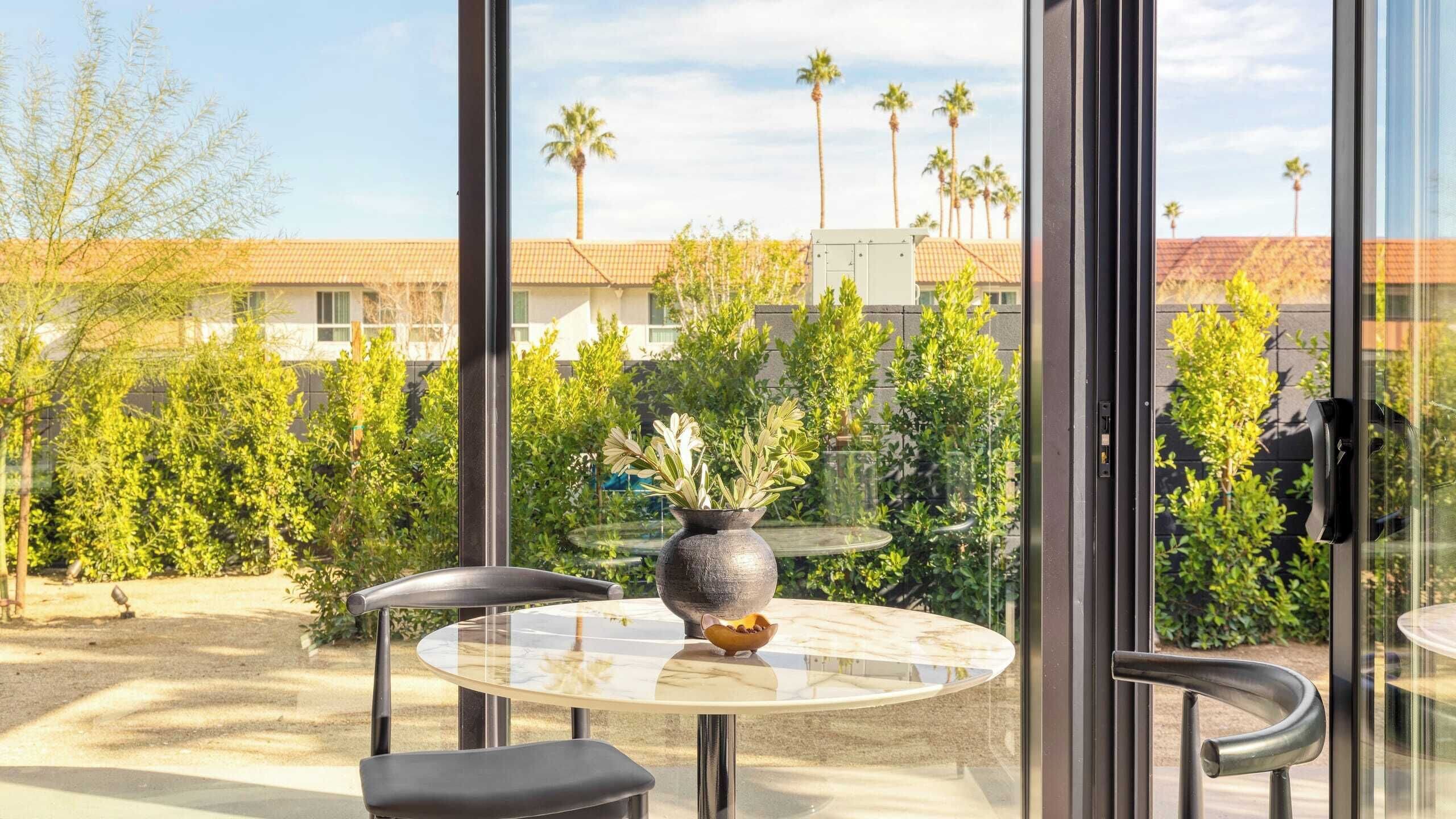 Un balcon avec une table en verre, deux chaises, un vase avec des plantes et une vue sur des palmiers et des immeubles.