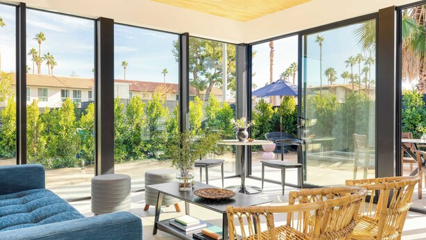 Een moderne woonkamer met grote glasramen, een zithoek buiten en uitzicht op palmbomen en huizen.