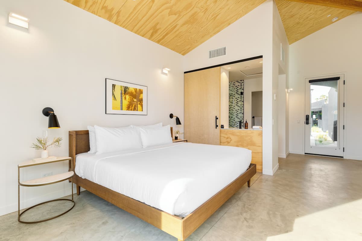 A modern bedroom with a wooden bed, white bedding, a bedside table with a lamp, a framed picture on the wall, and a sliding door leading to a balcony.