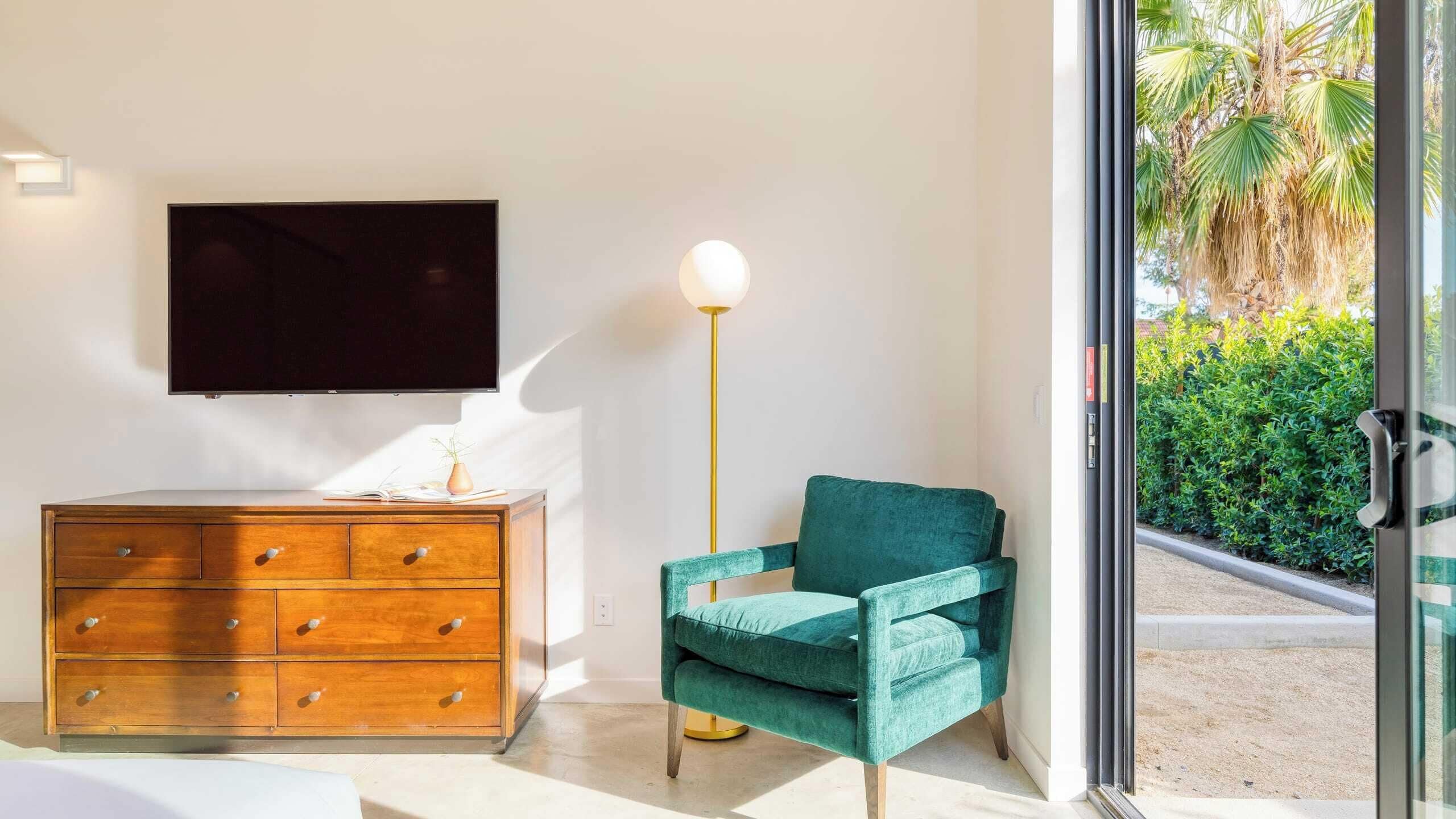 A modern living room with a wooden dresser, a teal armchair, a floor lamp, and a TV mounted on the wall.