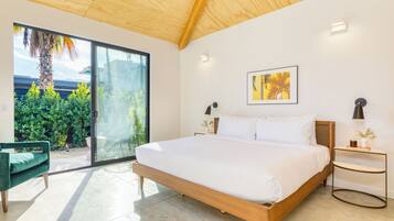 Ein modernes Schlafzimmer mit einem großen Bett, einem grünen Sessel, einem kleinen Tisch und Blick auf einen Garten durch eine Schiebetür.