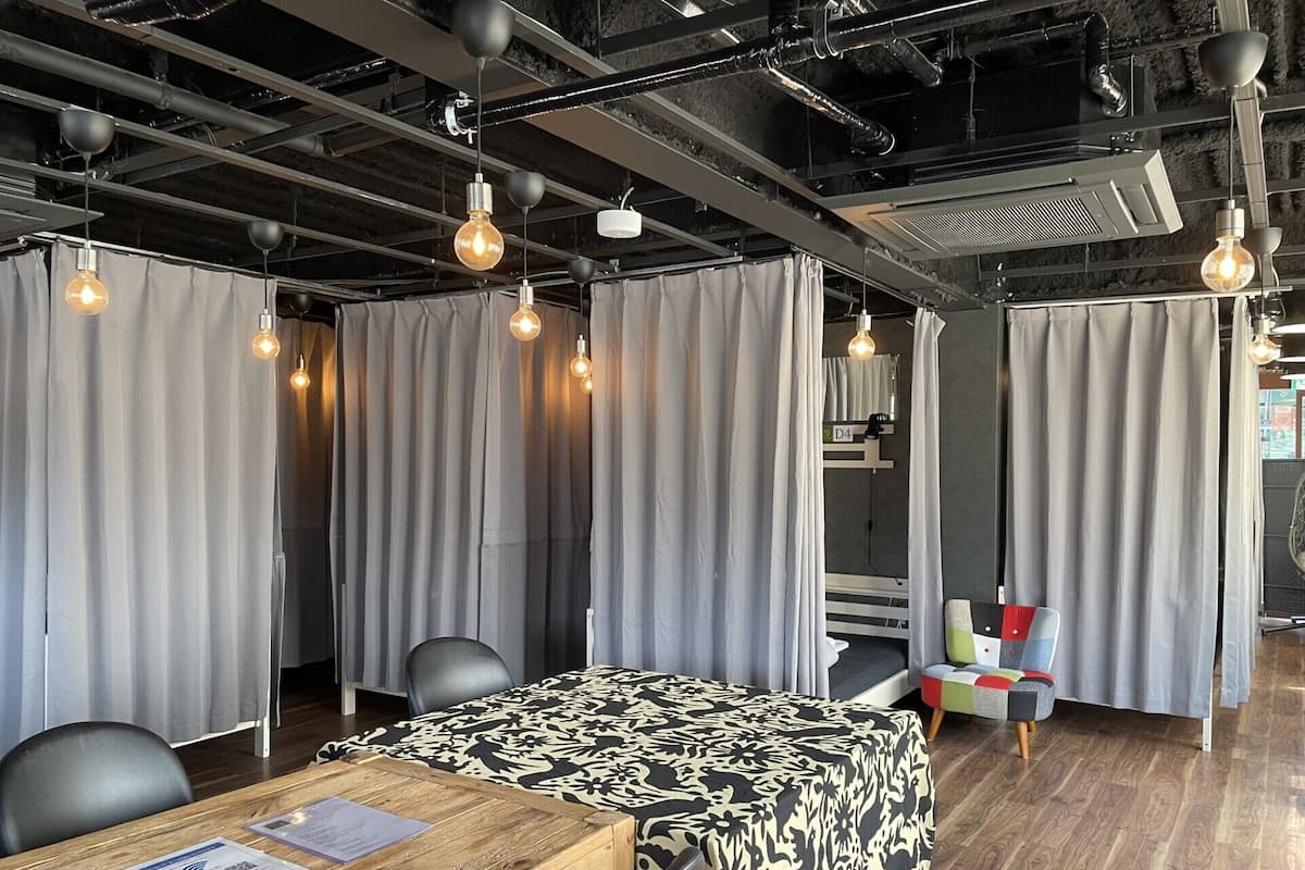 Habitación de hotel con una mesa de madera, una cama con colcha estampada, cortinas grises y lámparas colgantes.