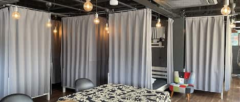 A hotel room with a wooden table, a bed with a patterned bedspread, grey curtains, and hanging pendant lights.