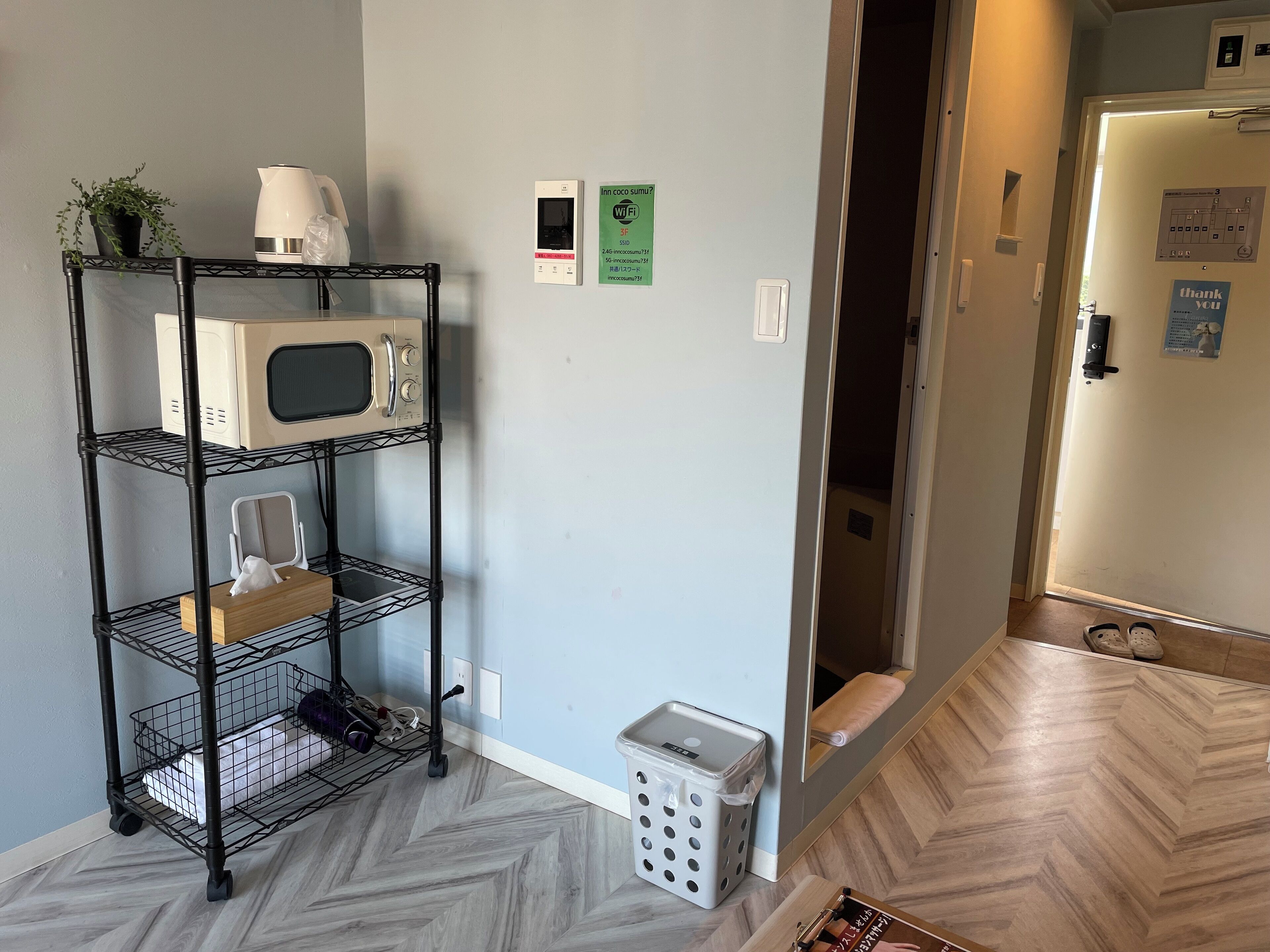 A kitchenette with a microwave, a coffee maker, and a shelf with a plant. There are two Wi-Fi signs on the wall and a door with a 'thank you' sign.
