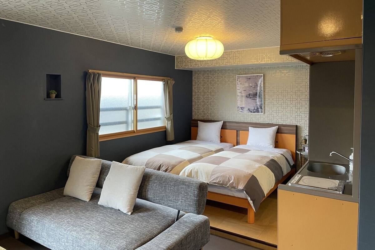 Habitación con dos camas, un sofá, una ventana con cortinas, una lámpara de pared y una zona de cocina.