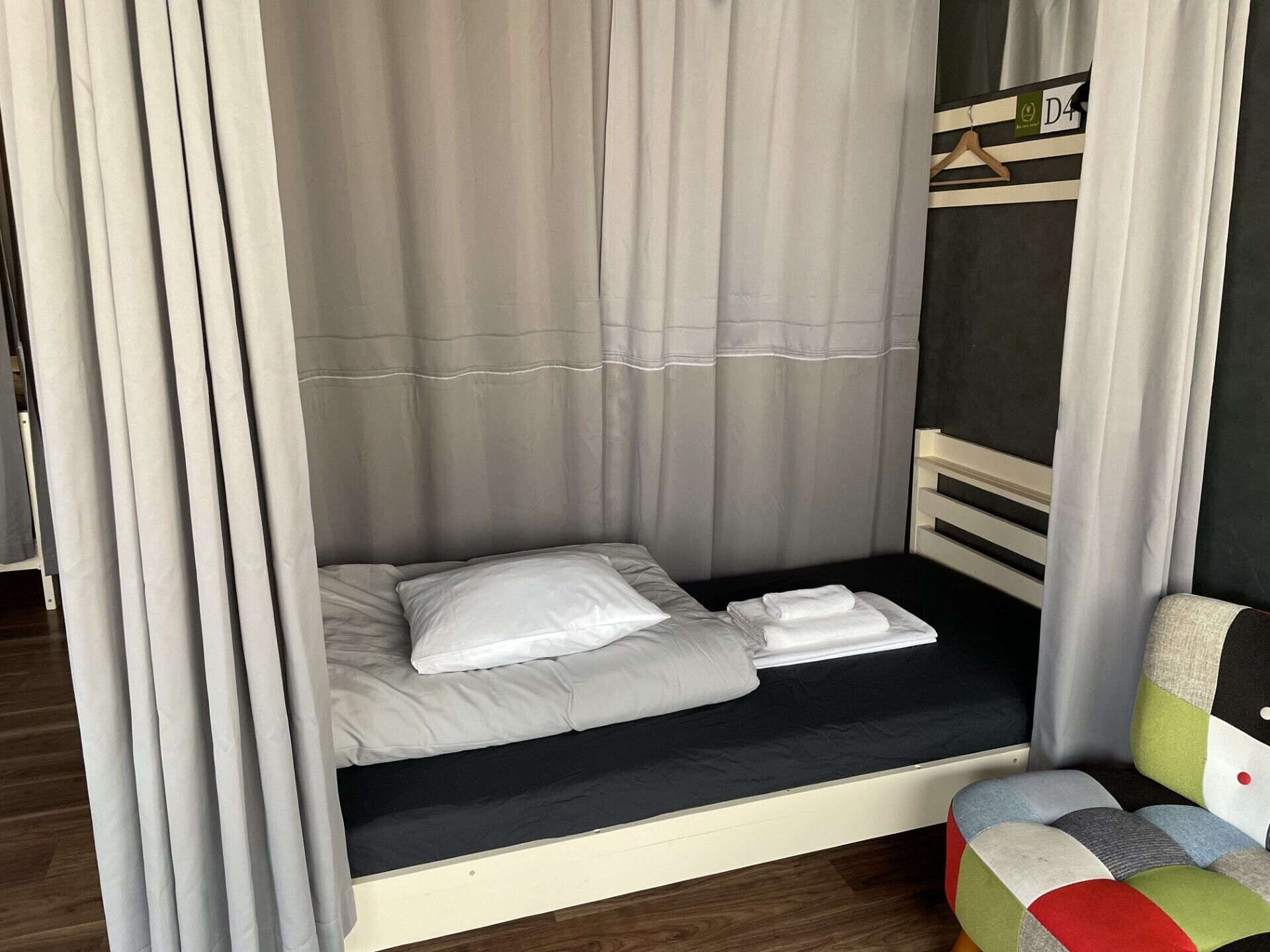 Una cama individual con colchón y almohadas blancos, cabecera de color oscuro y mesita de noche con un cojín colorido.