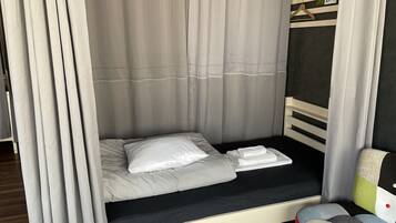 Una cama individual con colchón y almohadas blancos, cabecera de color oscuro y mesita de noche con un cojín colorido.