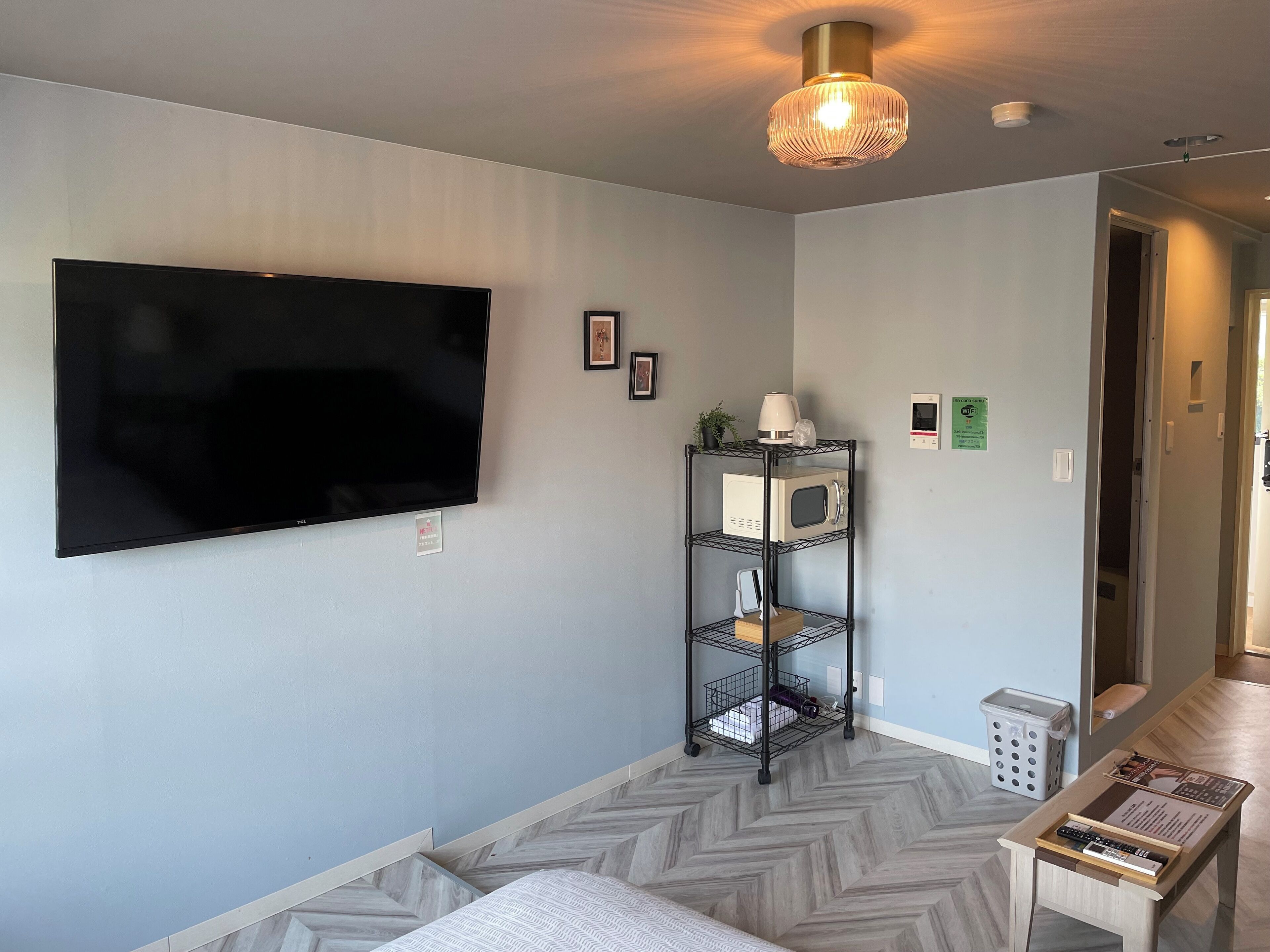 A modern living room with a flat-screen TV mounted on the wall, a black metal shelf with a microwave and other items, a patterned herringbone floor, and a small table with a remote control.