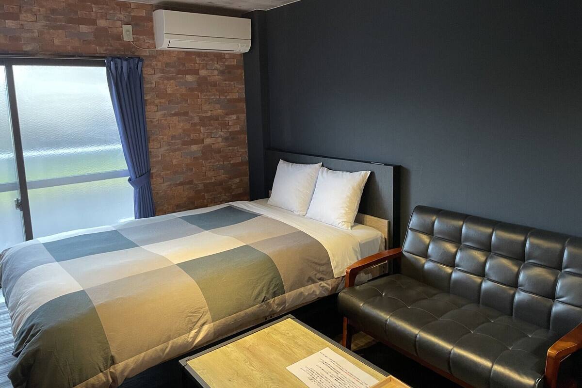Una habitación de hotel con cama, sofá, ventana con cortinas, pared de ladrillo y unidad de aire acondicionado.