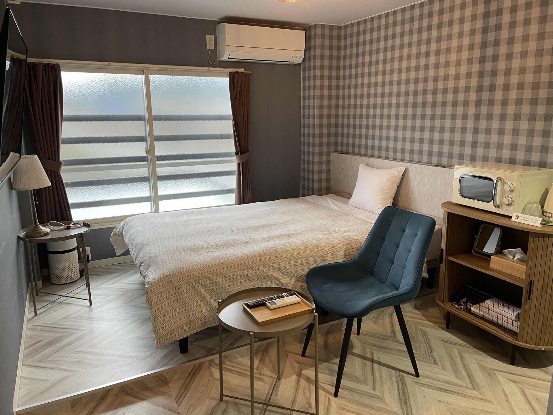 Hotelkamer met een bed, een stoel, een klein tafeltje, een magnetron, een prullenbak en een raam met gordijnen.