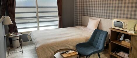 Hotelkamer met een bed, een stoel, een klein tafeltje, een magnetron, een prullenbak en een raam met gordijnen.