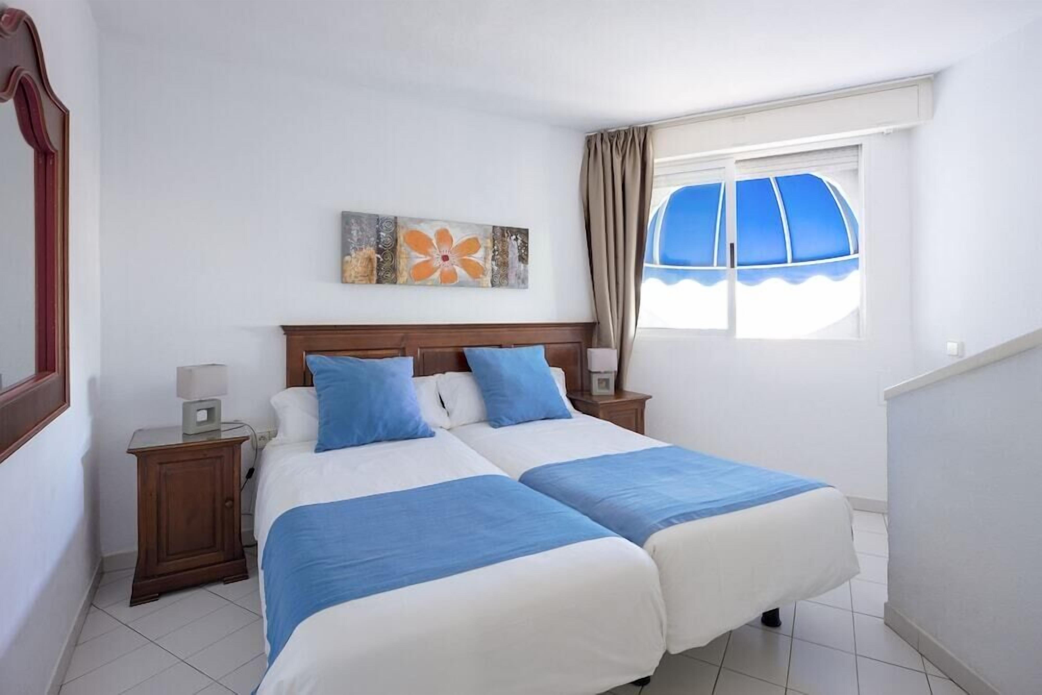 Cama doble con ropa de cama azul y blanca, cabecera de madera, mesita de noche con lámpara, un cuadro en la pared y una ventana con cortinas.