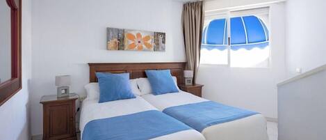 Cama doble con ropa de cama azul y blanca, cabecera de madera, mesita de noche con lámpara, un cuadro en la pared y una ventana con cortinas.