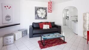 A modern living room with a grey sofa, red cushions, a glass coffee table, and a red rug. There is a kitchen visible through an archway. - Playa Flor Chill Out Bungalows (Arona)