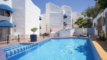 Una piscina con una característica de agua azul cristalina, rodeada de edificios blancos con detalles azules y una zona delimitada por una cerca.