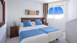 A double bed with blue and white bedding, a wooden headboard, a nightstand with a lamp, and a window with curtains. - Playa Flor Chill Out Bungalows (Arona)