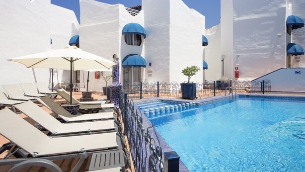 A poolside area with white buildings, blue umbrellas, and lounge chairs. - Playa Flor Chill Out Bungalows (Arona)