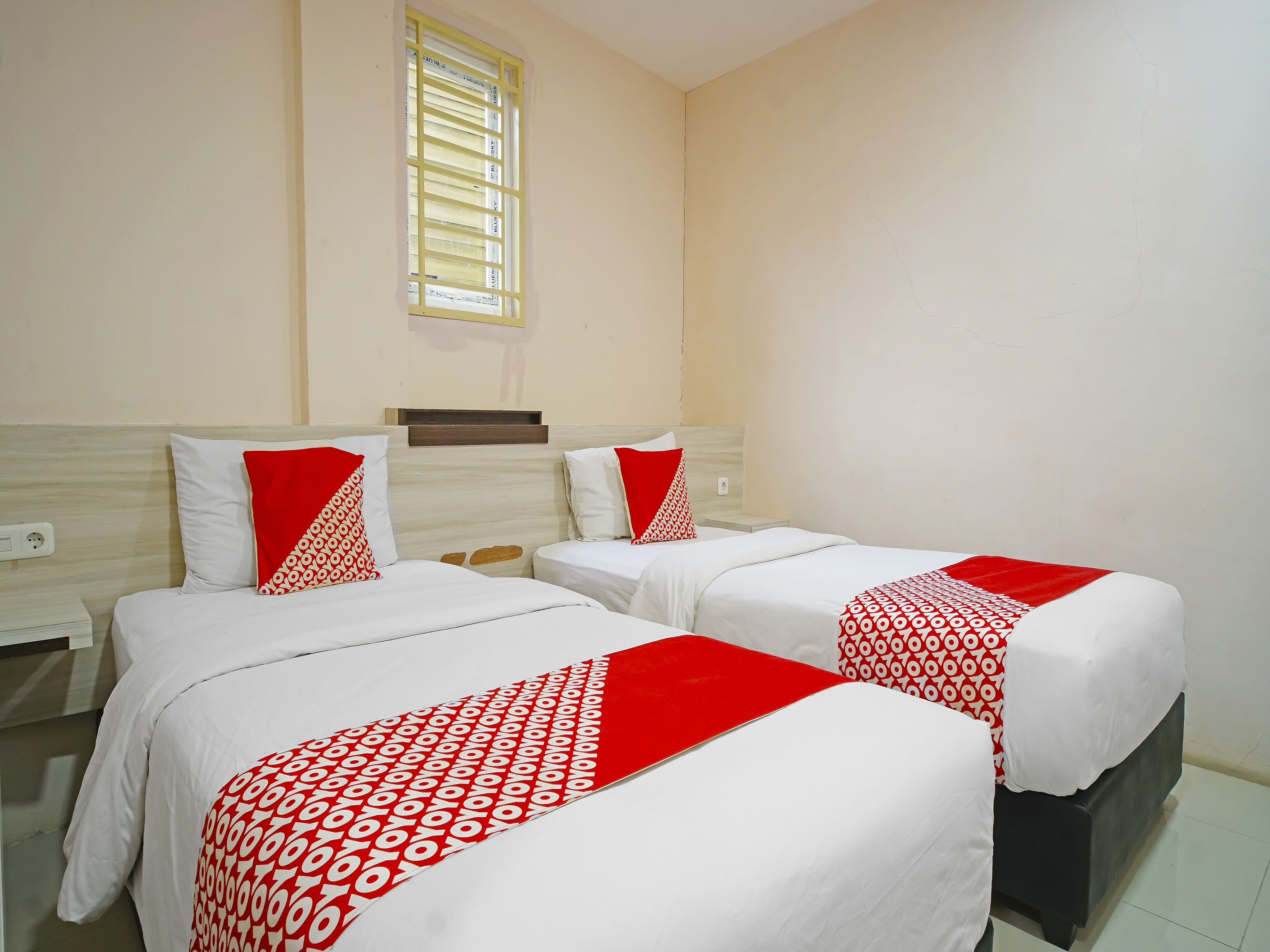 A hotel room with two beds, each with white bedding and red accents, a window with yellow blinds, and a plain wall.